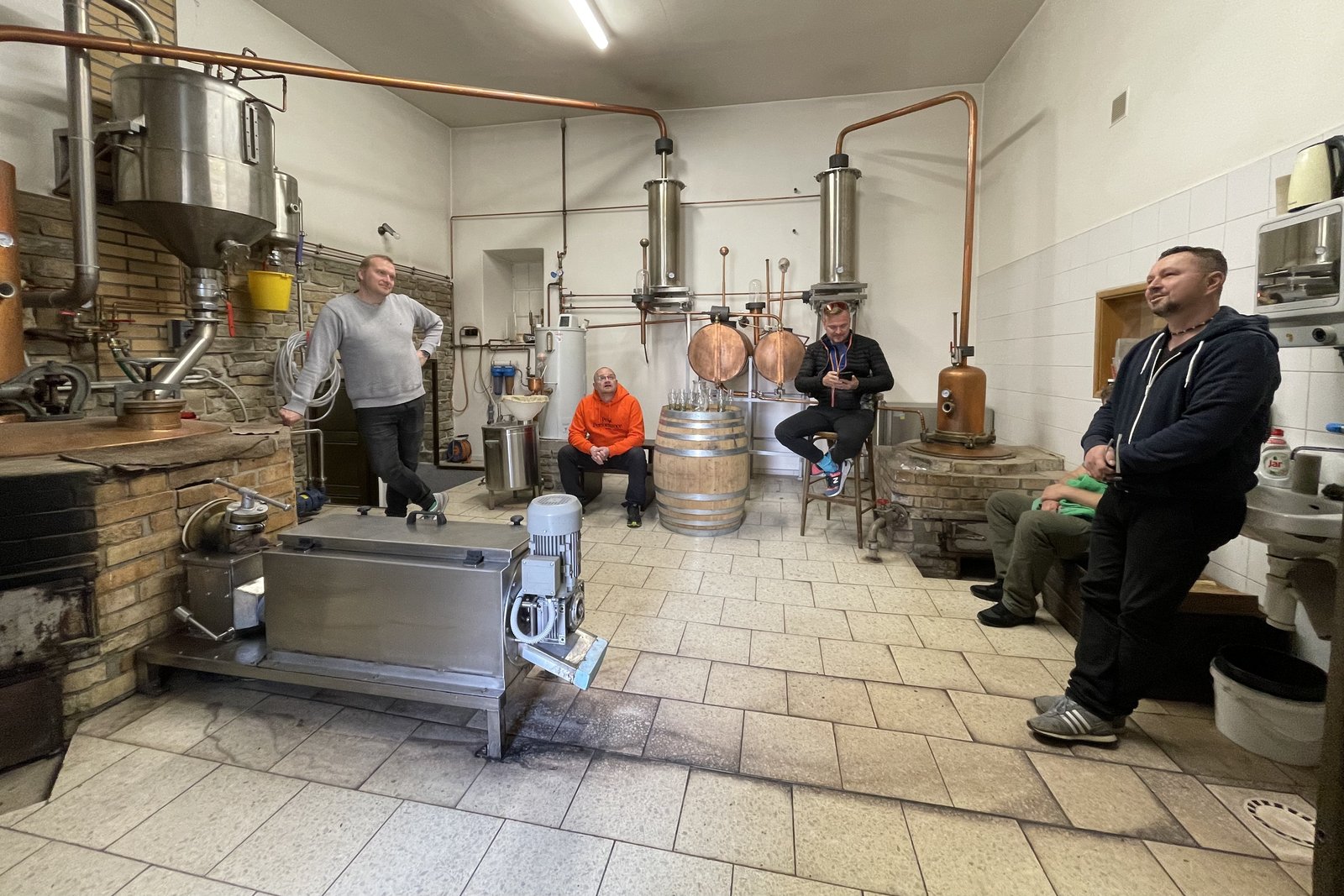 Inside the Kuželov distillery: copper stills, an oak barrel with tasting glasses, and a group listening to the distiller.